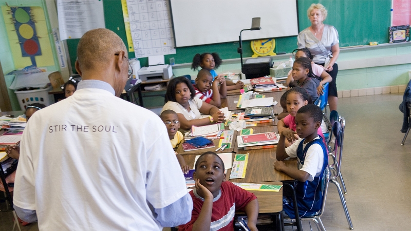 Classroom full of students, an adult in front of the room with the text