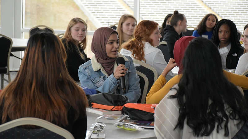Photo of a group of young women at tables, one speaking into a microphone at her seat